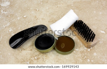 A set of black and brown shoe polish, soft application cloth, brush, and shoe horn on beige marbleized tile.