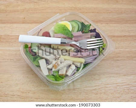An angle view of a prepared chef salad with fork in a closed plastic container on wood grain table.
