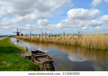 Typical landscape in Holland with an old boat in a ditch with a windmill, reeds and clouds.