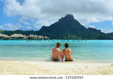 A honeymoon couple drinking a cocktail on the beach of a luxury vacation resort in the lagoon with a view on the tropical island of Bora Bora, near Tahiti, in French Polynesia.