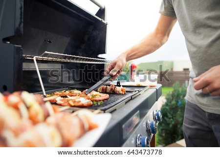 Hand of young man grilling some meat and vegetable-meat skewers on huge gas grill (Shallow DOF)