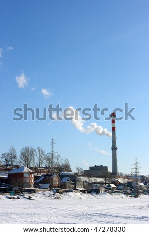 Urban landscape with a smoking stack and cottages on the river bank against a blue sky in winter