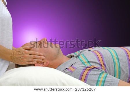 Female healer standing with hands gently positioned either side of supine male patient\'s temples channeling healing energy