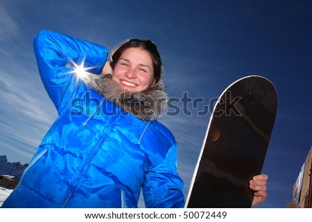 beautiful young woman standing on snow, smiling and holding a snowboard. against the sun