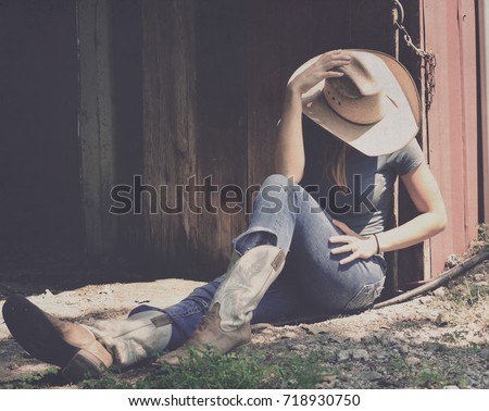 Girl in western wear relaxes in shadows of barn, representing agriculture industry in cowboy hat, jeans and cowboy boots.