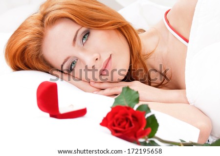 Beautiful girl on white bed. Flower and wedding ring in a gift box on a pillow next to her