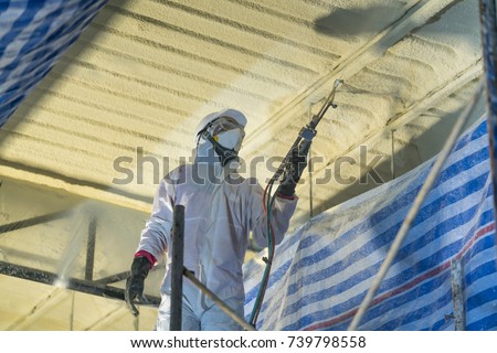 Technician spraying foam insulation using Plural Component Gun for polyurethane foam - Repair tool in the white protect suit applies a construction foam from the gun to the roof of a warehouse.