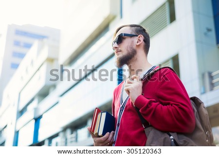 Caucasian man stands in front of hi-tech building and holds books and a backpack.