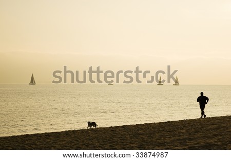 Silhouette of fit man and his dog exercise on the beach at sunset. Sailing boats on the water in the background