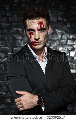 Waist up portrait of a handsome man wearing a suit, looking at the camera with zombie bloody make up on his face, against black brick wall