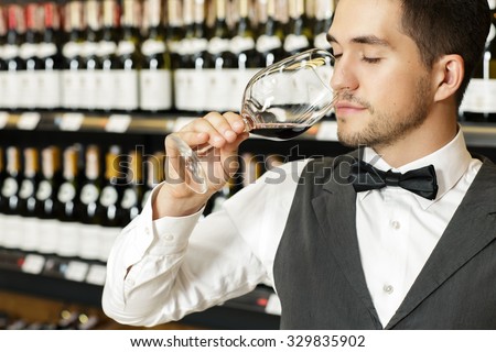 Portrait of a handsome bearded sommelier smelling red wine from the large glass in wine store, wine shelves on the background
