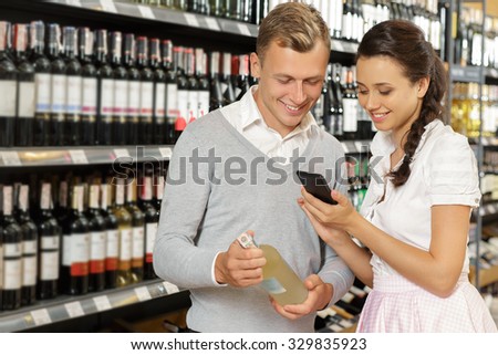 Making a decision. Young smiling couple standing in a supermarket holding a bottle of wine and searching some information about it on the internet using smartphone