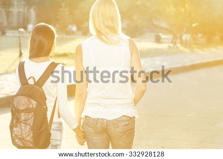 Walking side by side. Horizontal shot from behind of mother walking her teen daughter with a backpack to school holding hands