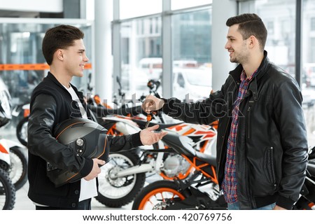 Congrats on your new ride! Portrait of a handsome young biker man in a leather jacket passing motorbike keys to a motorcycle salon customer