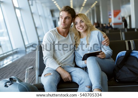 Travelling together. Beautiful young couple in love embracing sitting at the waiting area of the airport terminal waiting for boarding smiling looking away love family honeymoon travel airport concept
