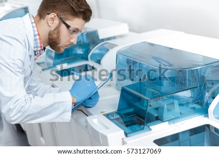 Making notes. Professional male scientist making notes on his clipboard watching analyzing machine during his research at the modern laboratory professionalism experiment science medicine concept