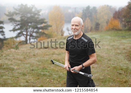 People, age, trekking, sports, adventure and hobby concept. Outdoor shot of unshaven sporty man pensioner with gray hair having excited happy look while exercising outdoors with nordic walking poles