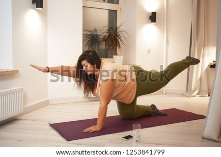 Sid view of overweight obese young woman wearing t-shirt and leggings doing physical training on mat to strengthen legs, arms, abs and spine. Weght loss, fitness, sports and active lifestyle concept