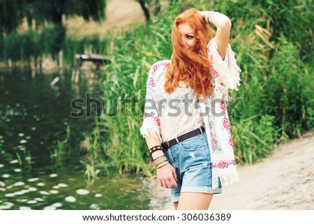 Bohemian fashion girl in kimono and blue denim shorts. Red haired young woman in summer outfit. Portrait of a beautiful girl standing by the river, enjoying a warm summer day in the nature.