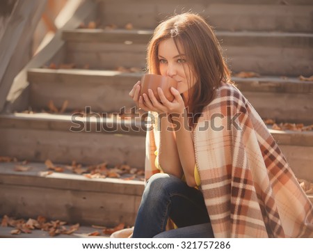 Beautiful girl resting and drinking tea sitting in autumn garden on the steps, wrapped in a woolen plaid blanket.