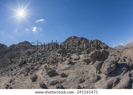 Peak of rocky mountain in arid desert landscape against blue sky background with sun
