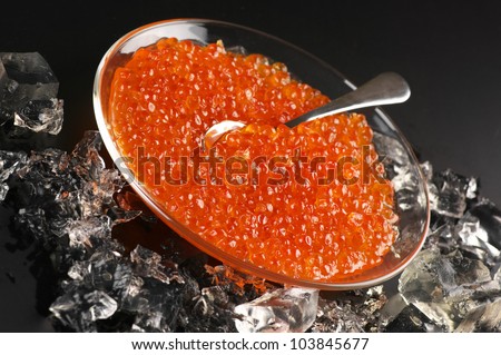 Salmon caviar in glass plate with spoon and ice on black background.