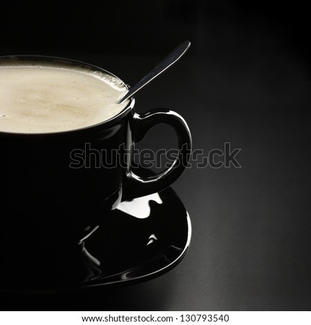 Black cup of coffee with cream and spoon close-up on dark background.