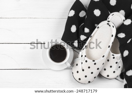 Pair of feminine slippers with white and black polka dot decor, woolen knitwear and cup of coffee on white wood.