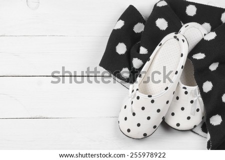 Pair of feminine slippers with white and black polka dot decor and woolen scarf on white wooden floor.