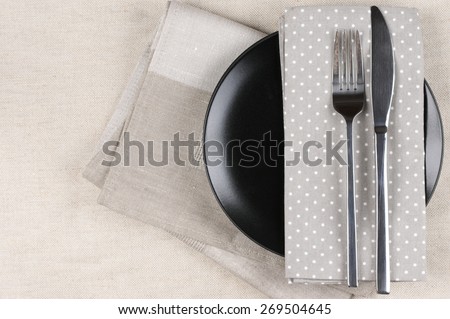 Table setting: black plate, fork and knife with napkins on linen tablecloth. Top view point.