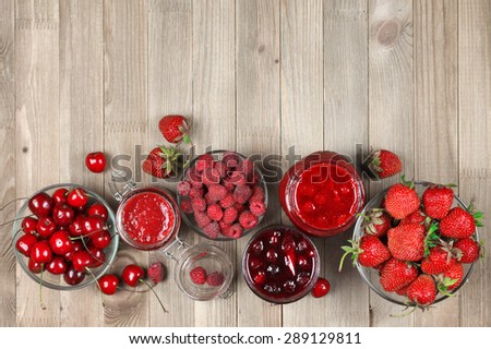 Assorted jams in glass jars and fresh berries in bowls on rustic wooden background. Top view point.
