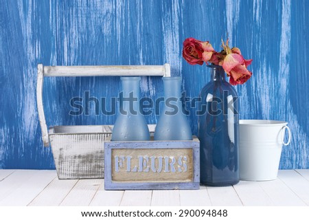 Home interior details: dried roses in blue glass bottle, rustic wooden boxes and white bucket against blue painted wall.