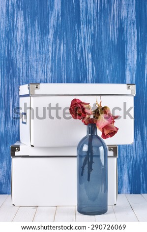 Home interior details: dried roses in blue glass bottle and white boxes against blue painted wall.