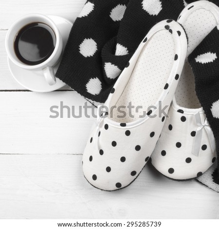 Pair of feminine slippers with white and black polka dot decor, woolen knitwear and cup of coffee on white wood.