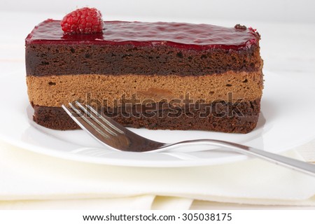 Slice of chocolate layered cake with souffle, fruity jelly and raspberry on white plate.