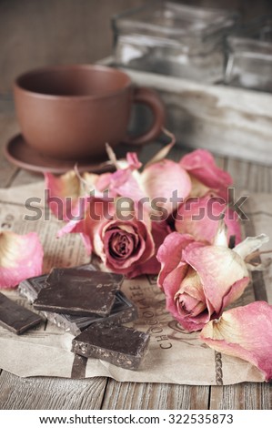 Slices of dark chocolate on rough paper, dried pink roses heads and coffee cup on wooden table. Shallow DOF, focus on chocolate.