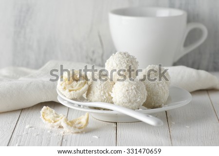 White whole and broken coconut candy balls in plate and cup on rustic wooden background. White food styling.