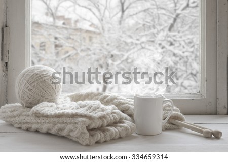 Cozy winter still life: mug of hot tea and warm woolen knitting on vintage windowsill against snow landscape from outside.
