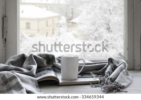 Cozy winter still life: mug of hot tea and opened book with warm plaid on vintage windowsill against snow landscape from outside.