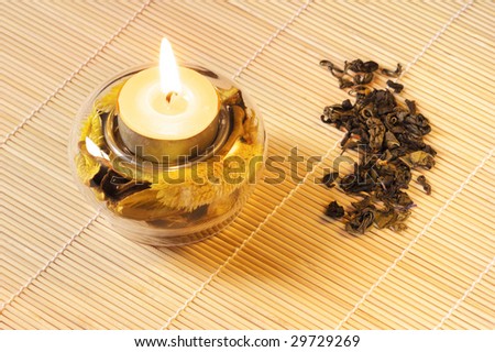 Burning scented candle with dried flowers and green tea on wood mat.