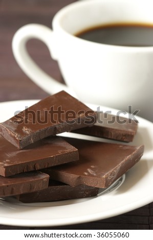 Dark chocolate close-up on white plate and white cup of coffee on brown background.