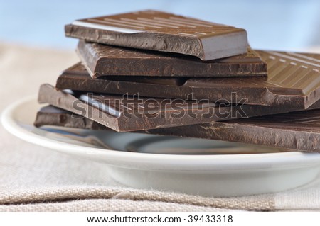 Stack of broken dark chocolate bar in glass saucer on beige napkin against blue background.