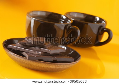 Stack of broken dark chocolate bar and two brown cups of coffee on yellow background.
