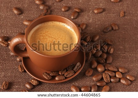 Brown ceramic cup of coffee with froth and coffee beans on brown canvas.