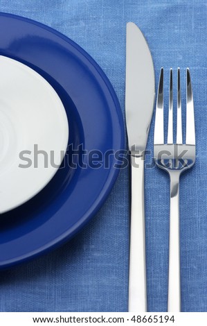 Blue and white plates, stainless fork and knife on blue linen tablecloth.