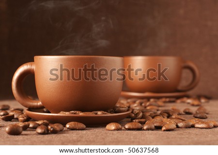Two brown ceramic cups of coffee with steam and coffee beans on brown canvas.
