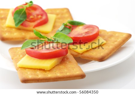 Three square crackers with slices of cheese, tomato and basil in white plate on white background.