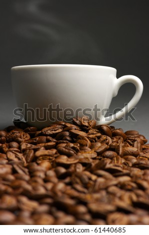 White cup of hot coffee on heap of roasted coffee beans against dark gray background.