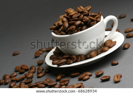 Roasted coffee beans in white cup with saucer on dark gray background.