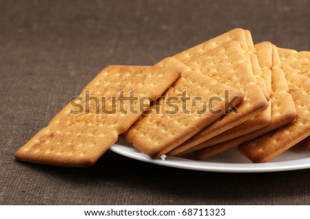 Heap of square crackers in white plate on brown canvas.